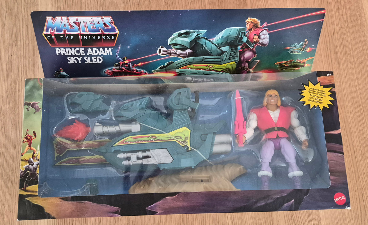 Masters of the Universe Prince Adam Sky Sled action figure in its original window box packaging, displayed on a light wood surface. The package includes the toy figure, the teal and yellow sky sled, and the illustrated header card.