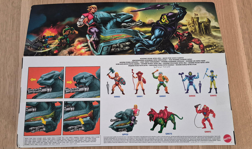 The back of a He-Man action figure box or booklet laid flat on a light wood surface, showing large comic-style artwork across the top and character details and names across the bottom. A red Mattel logo is visible in the lower right corner.