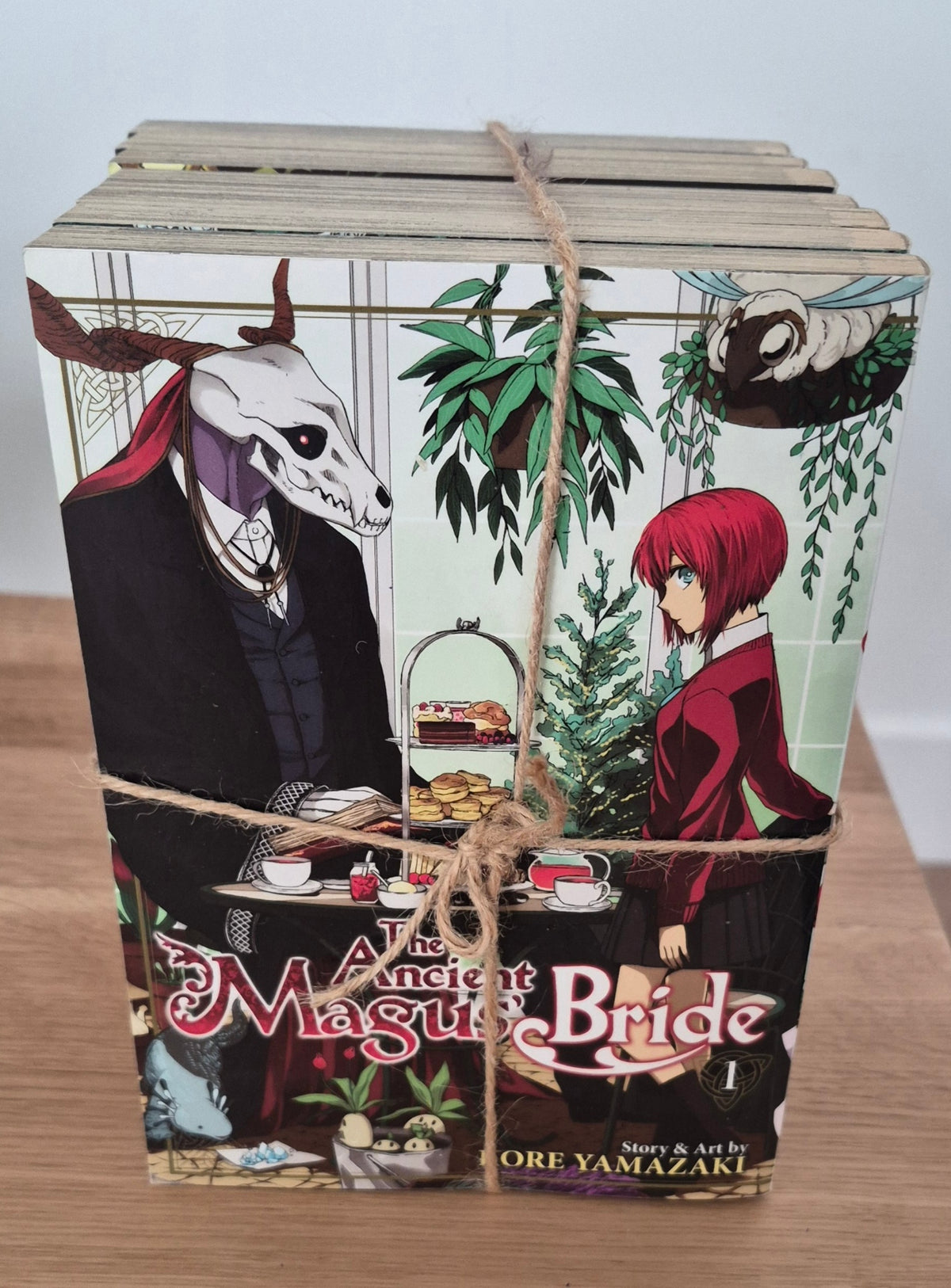A boxed set of The Ancient Magus Bride Volume 1 manga books tied with twine, placed on a wooden surface against a light gray wall.