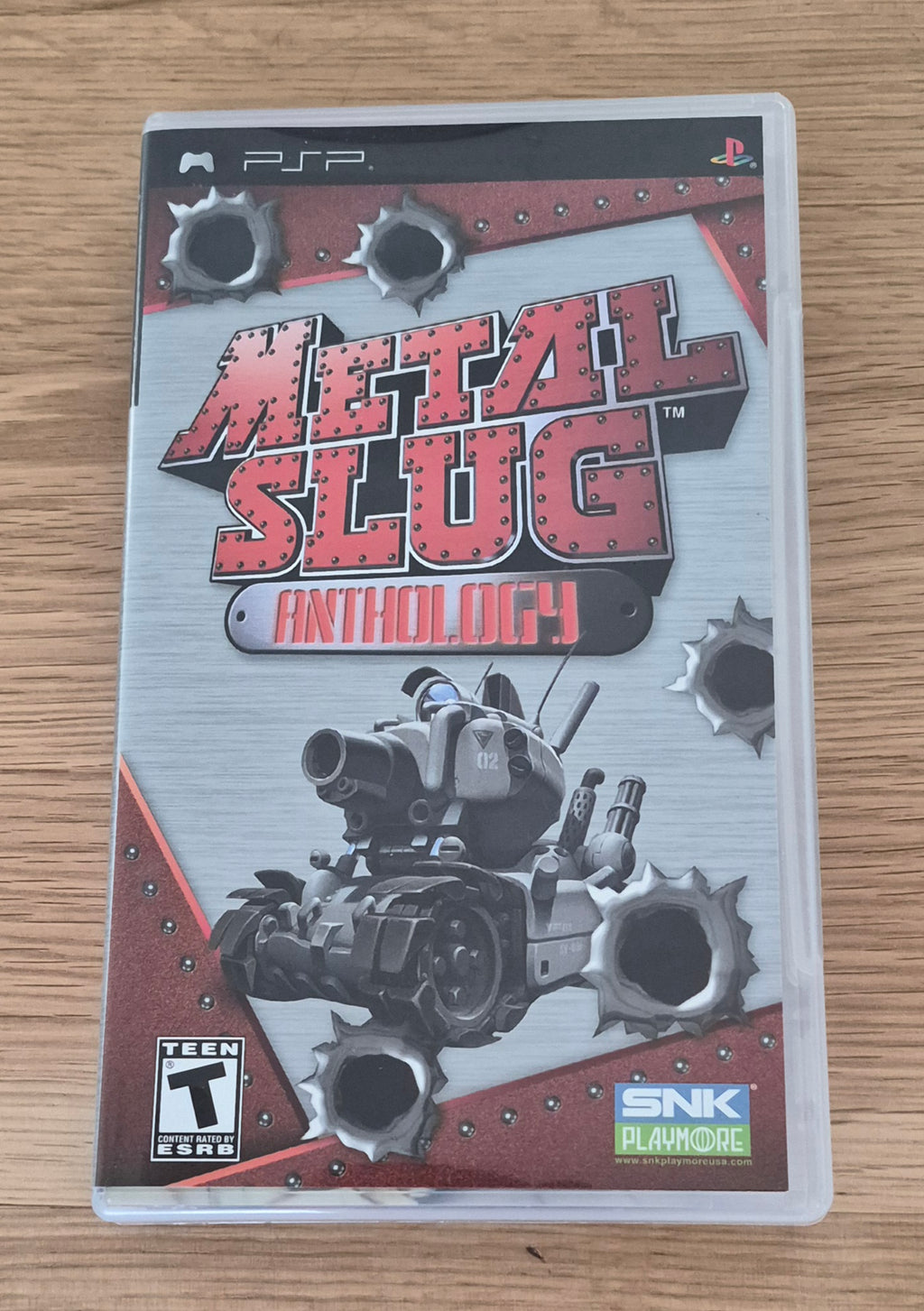 Metal Slug Anthology video game case for the Sony PSP, featuring the game's cover art with a tank and bullet holes, resting on a light wood surface.