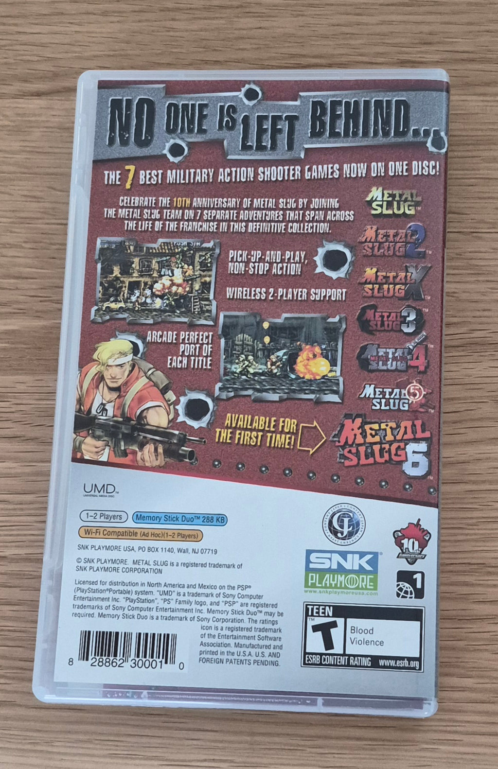 Back cover of the Metal Slug Anthology video game case, photographed from above on a light wood surface. The case shows the title, game artwork, feature callouts, and publisher logos including UMD and SNK.