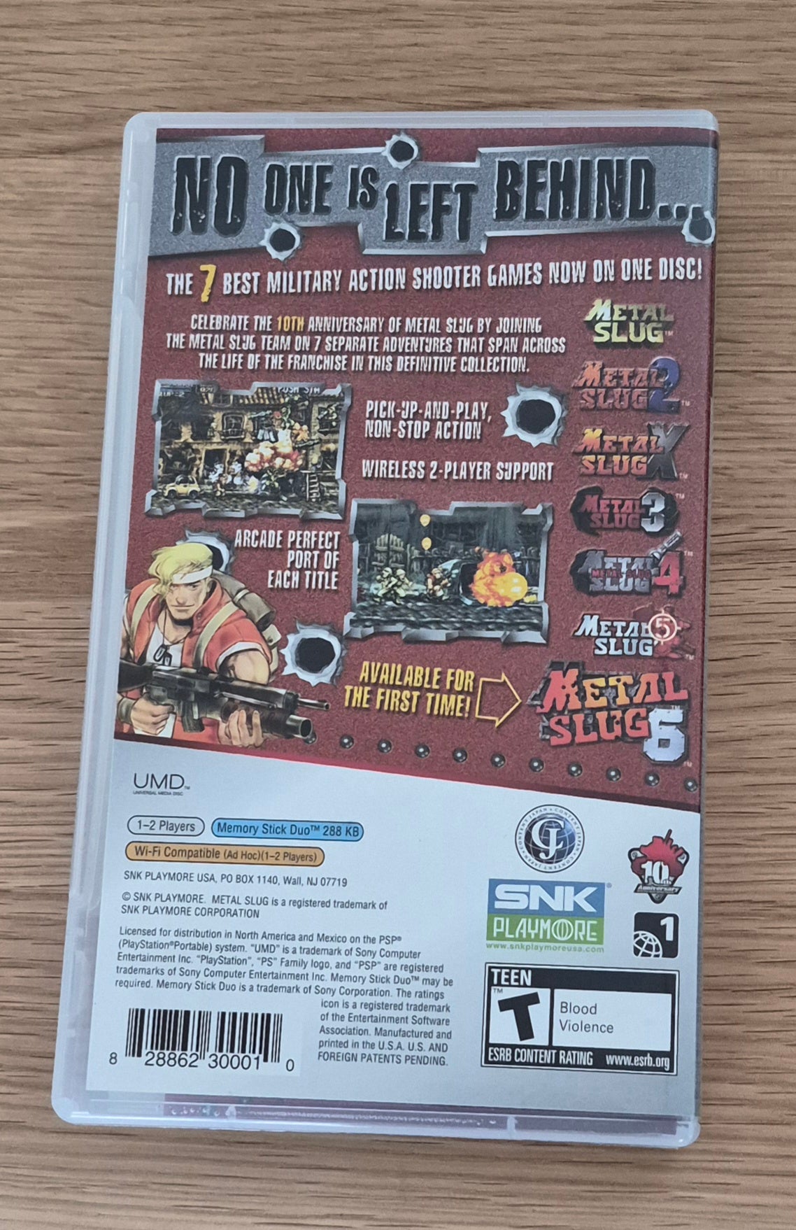 Back cover of the Metal Slug Anthology video game case, photographed from above on a light wood surface. The case shows the title, game artwork, feature callouts, and publisher logos including UMD and SNK.