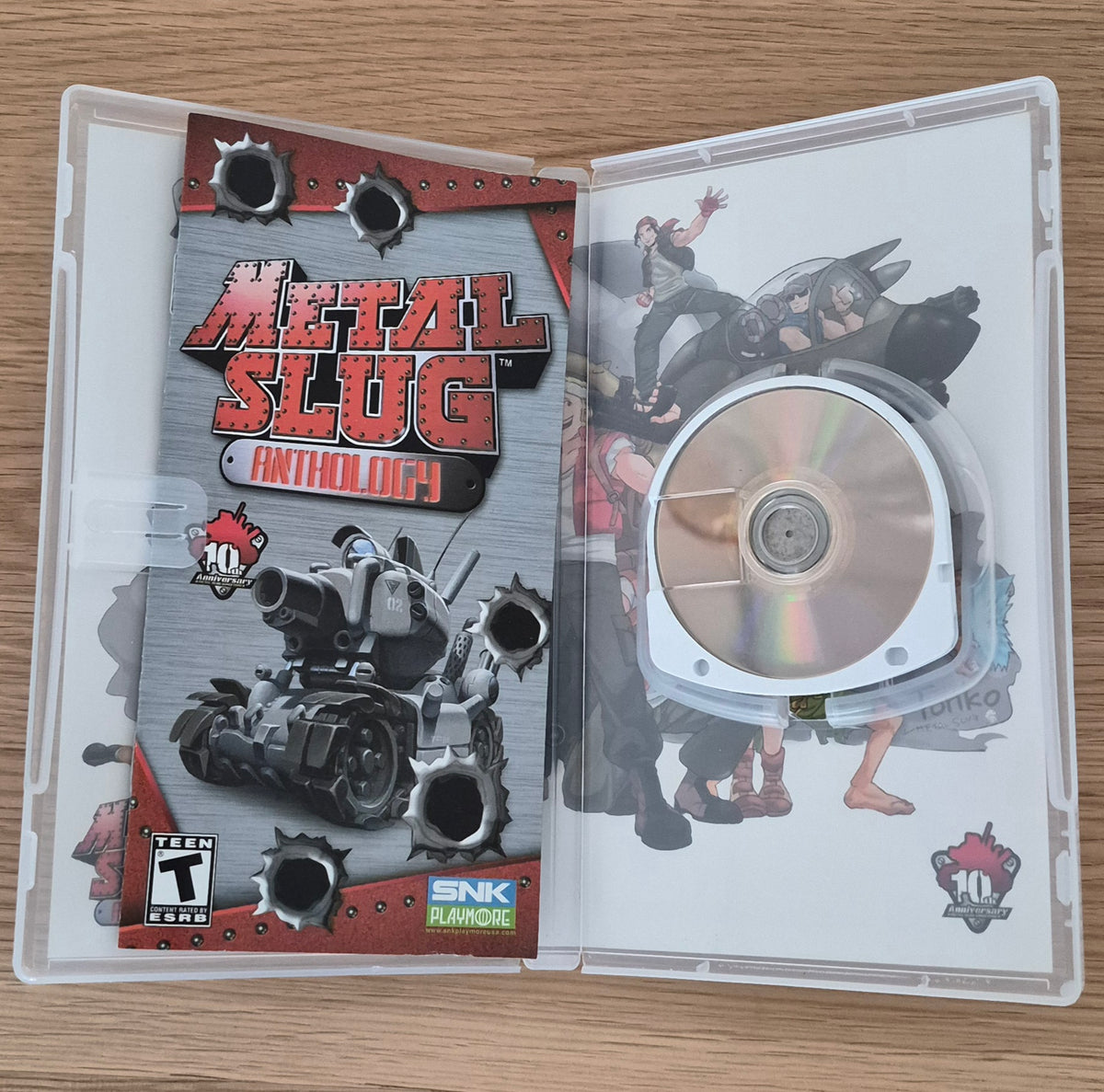 Metal Slug Anthology 10th Anniversary game case opened flat on a wooden surface, showing the printed cover art, a disc inside, and the SNK Playstation branding.